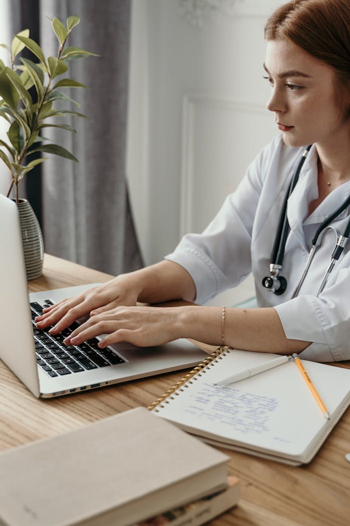 home-hero A professional female doctor typing on a laptop in her office with medical notes and stethoscope.