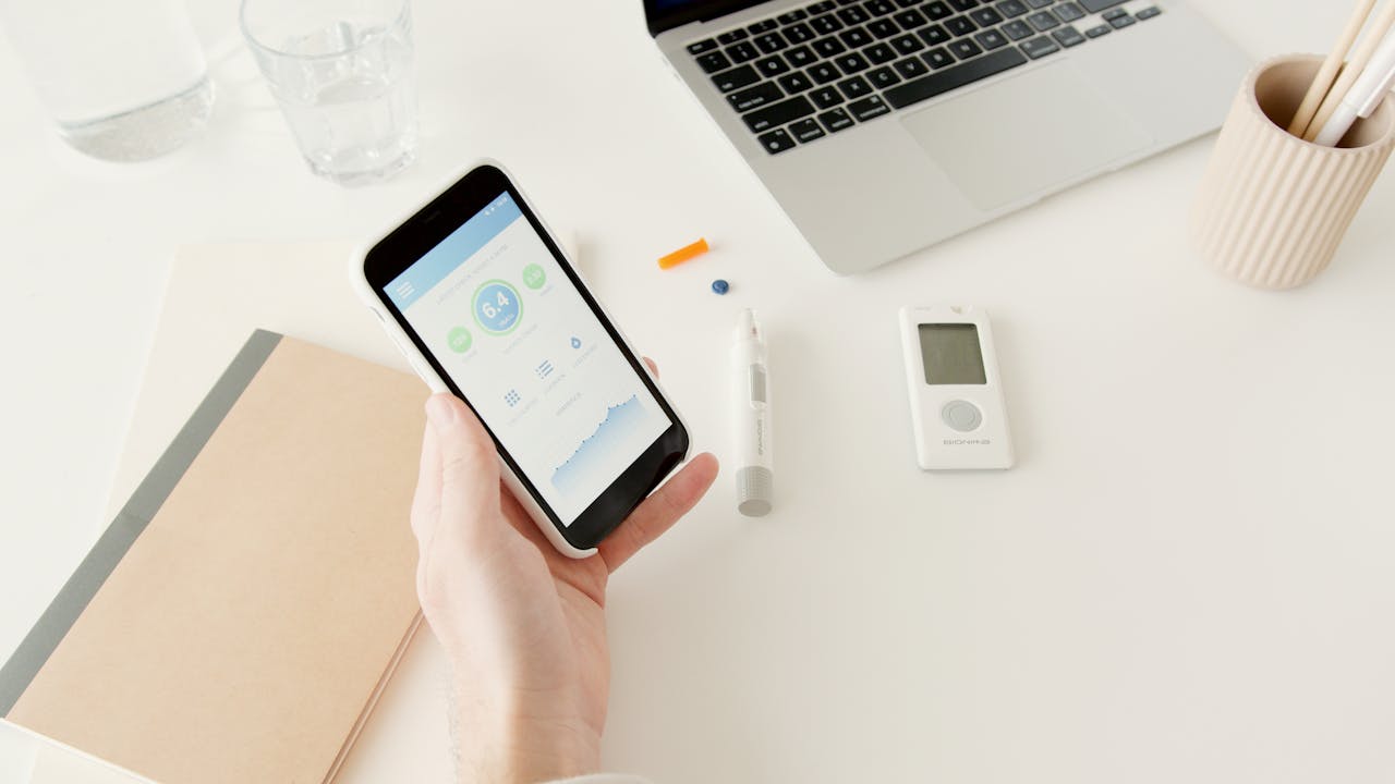who-we-are A person checks glucose levels on a smartphone app next to diabetes monitoring equipment.